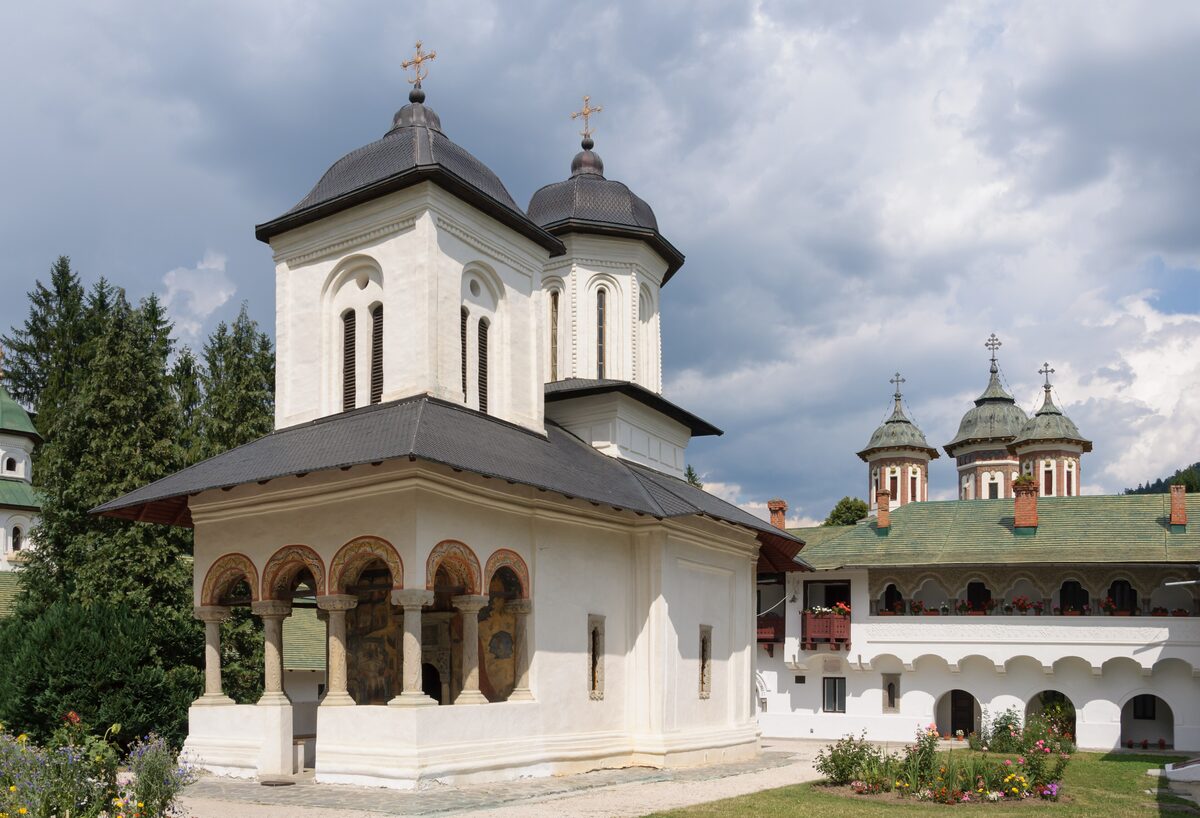 The small white old church of Sinaia Monastery with a painted porch, and the domes of the larger church behind