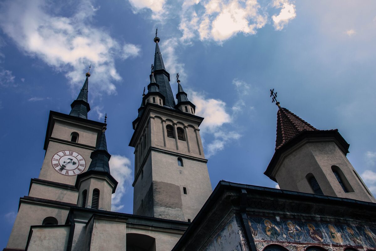 The Gothic spires and clock tower of St. Nicholas Church in Șchei, Brașov, against a blue sky with white clouds