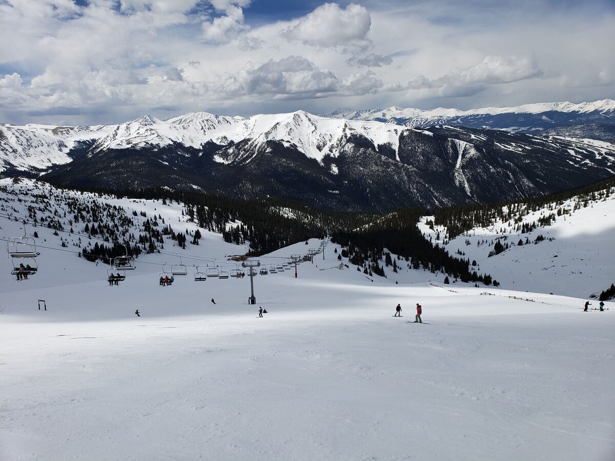 Skiers on a wide snowy slope in Montezuma Bowl at Arapahoe Basin, with a chairlift and snow-covered peaks behind