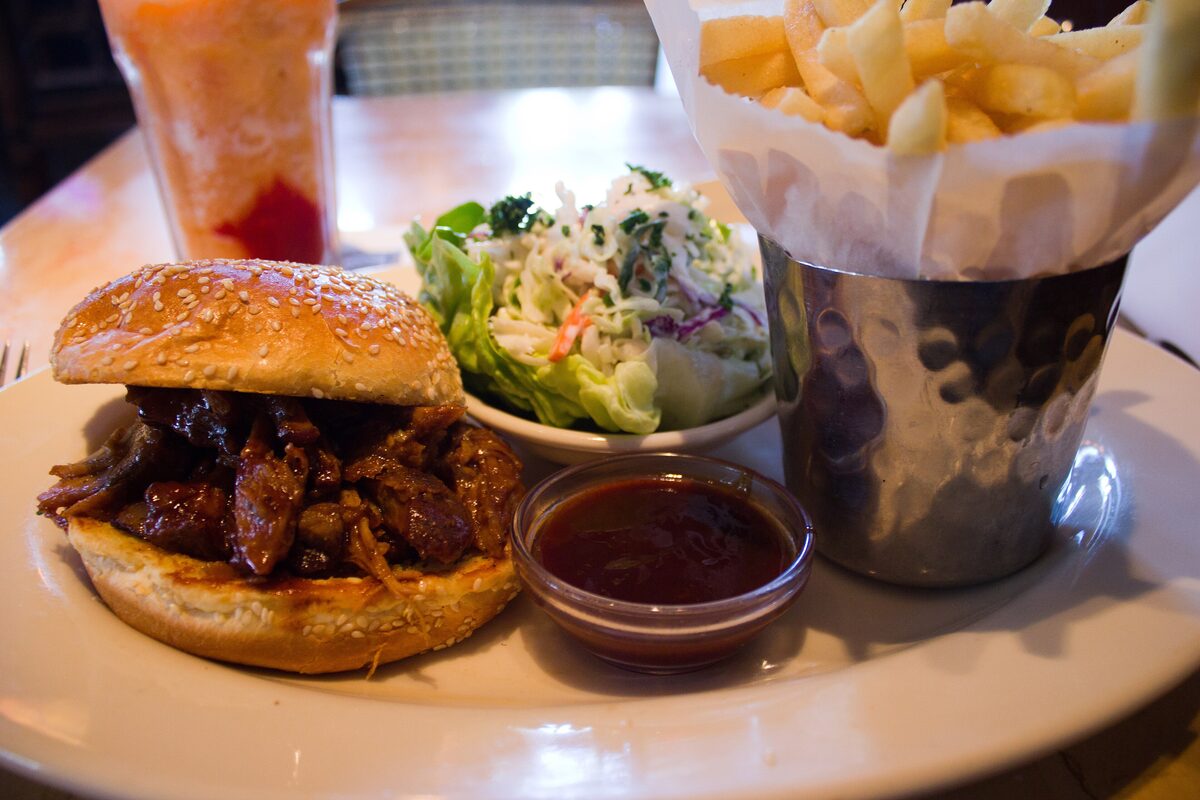 A pulled pork sandwich in a sesame bun with barbecue sauce, coleslaw and a cup of fries on a white plate