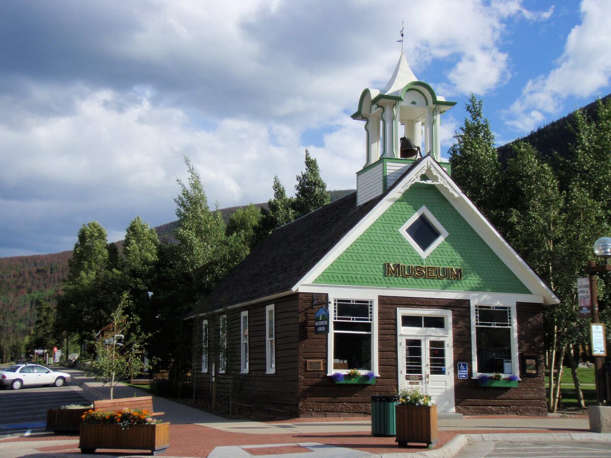 The old Frisco Schoolhouse with its green cupola and a Museum sign, with trees and mountain slopes behind