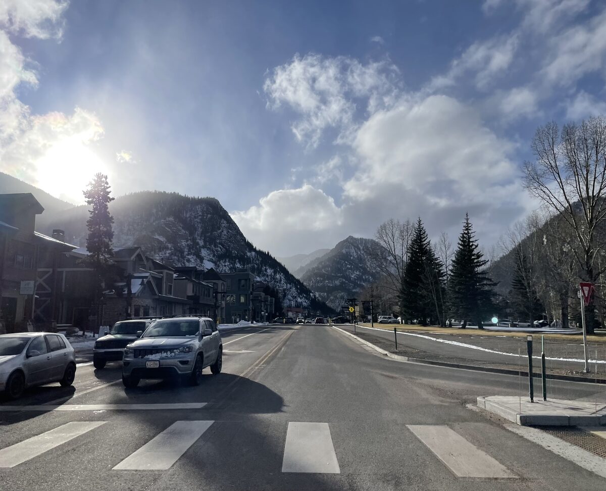 A sunlit street in Frisco with a pedestrian crossing, buildings on the left and mountains rising behind the town