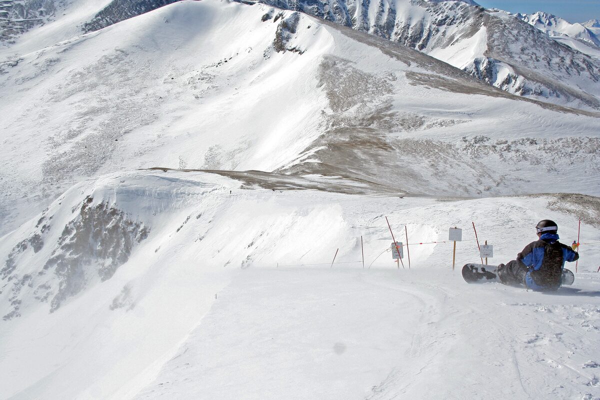 A high, wind-scoured ridge of the Imperial terrain above treeline, with a snowboarder resting at the edge and the Tenmile Range beyond