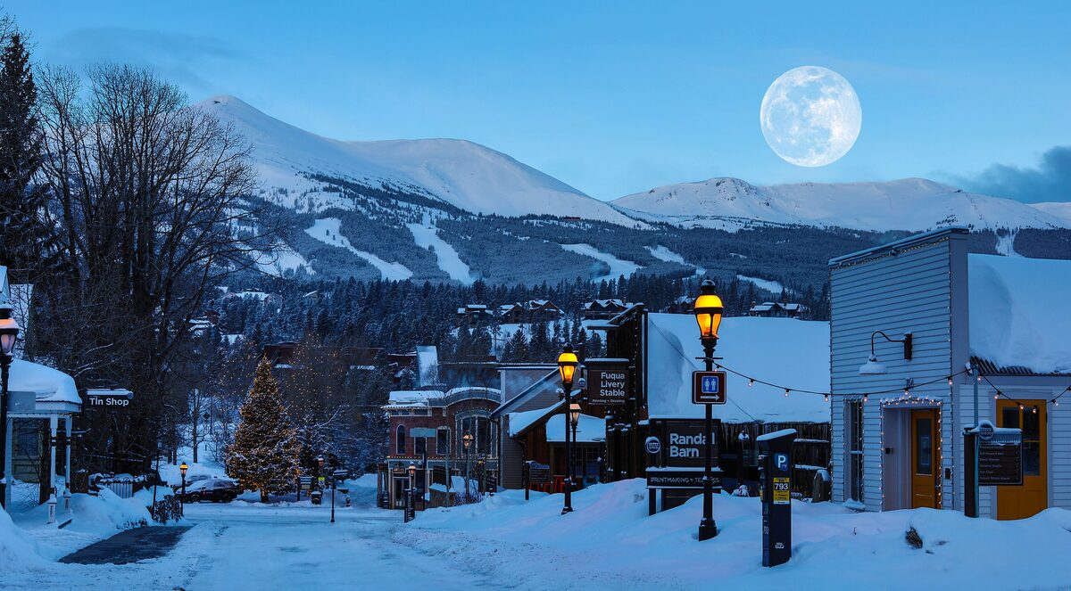 Breckenridge's snowy Main Street at dusk with street lamps, a Christmas tree and a full moon over the ski slopes