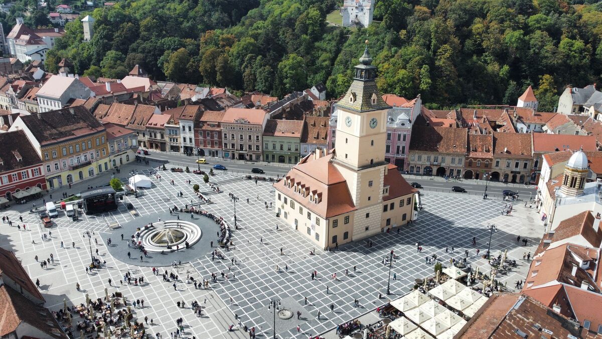 Aerial view of Brașov's Council Square, with the old yellow Council House and its clock tower in the middle of the paved square, below a forested hill
