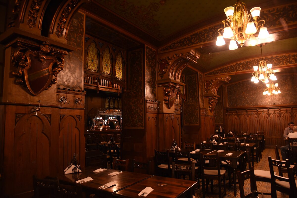 The dark carved-wood dining hall of Caru' cu Bere, with heraldic shields, stained-glass panels, brass chandeliers and set wooden tables