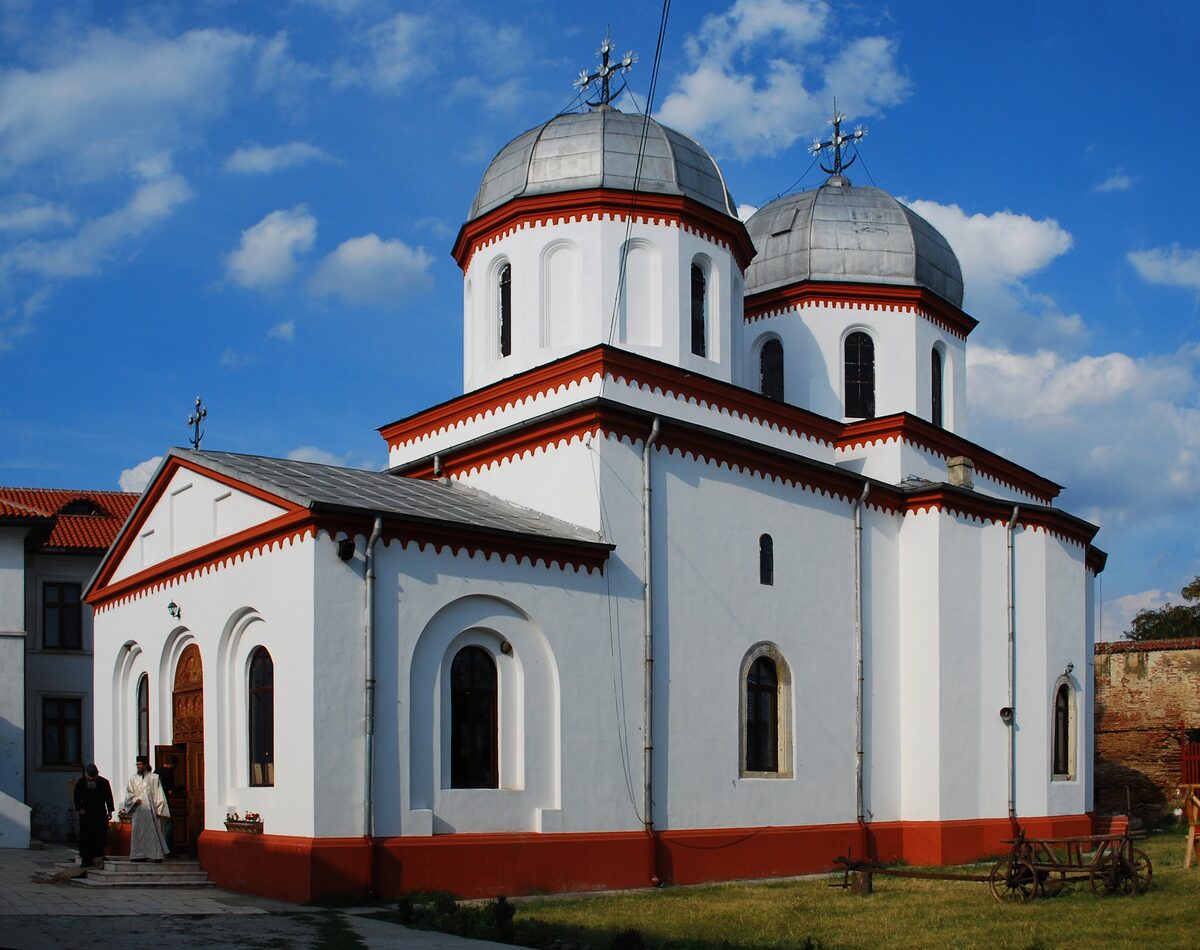 The white church of Comana Monastery with red trim and two silver domes, against a blue sky with clouds