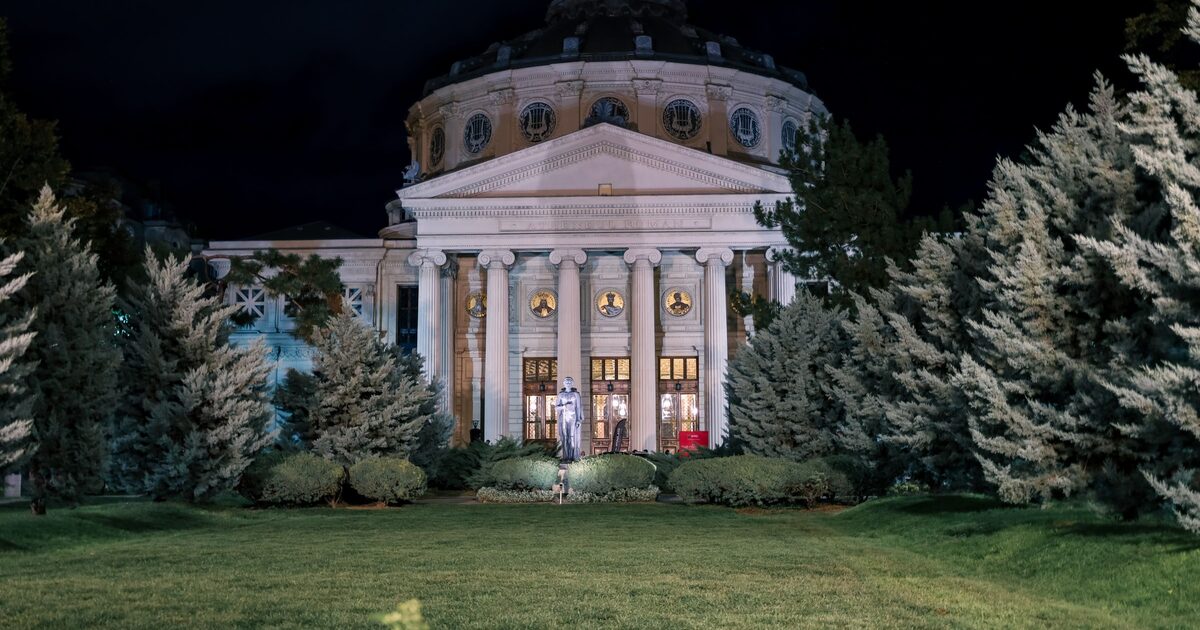 The Romanian Athenaeum lit up at night, its domed rotunda and columned portico behind a lawn and fir trees, with the statue of Mihai Eminescu in front