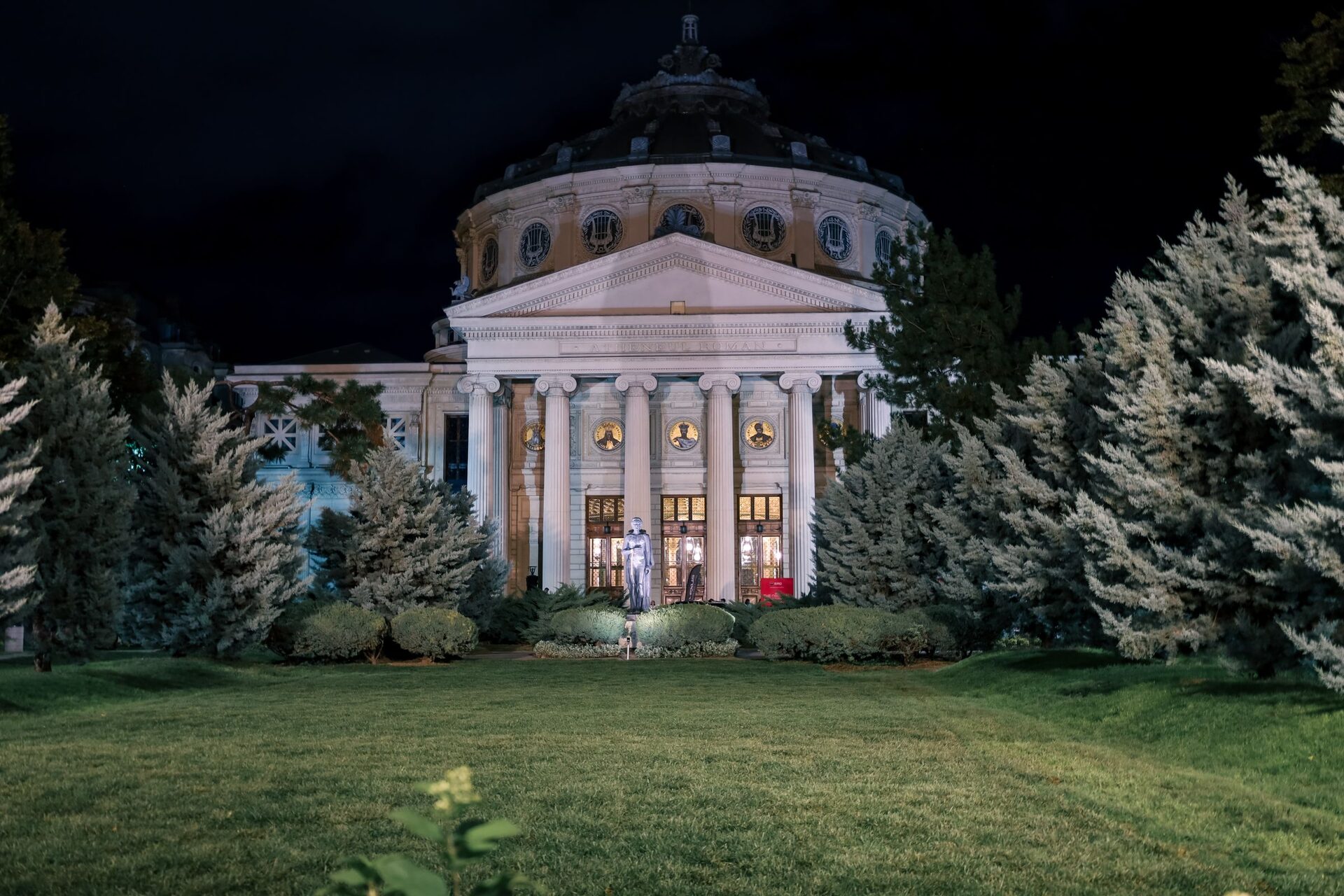 The Romanian Athenaeum lit up at night, its domed rotunda and columned portico behind a lawn and fir trees, with the statue of Mihai Eminescu in front