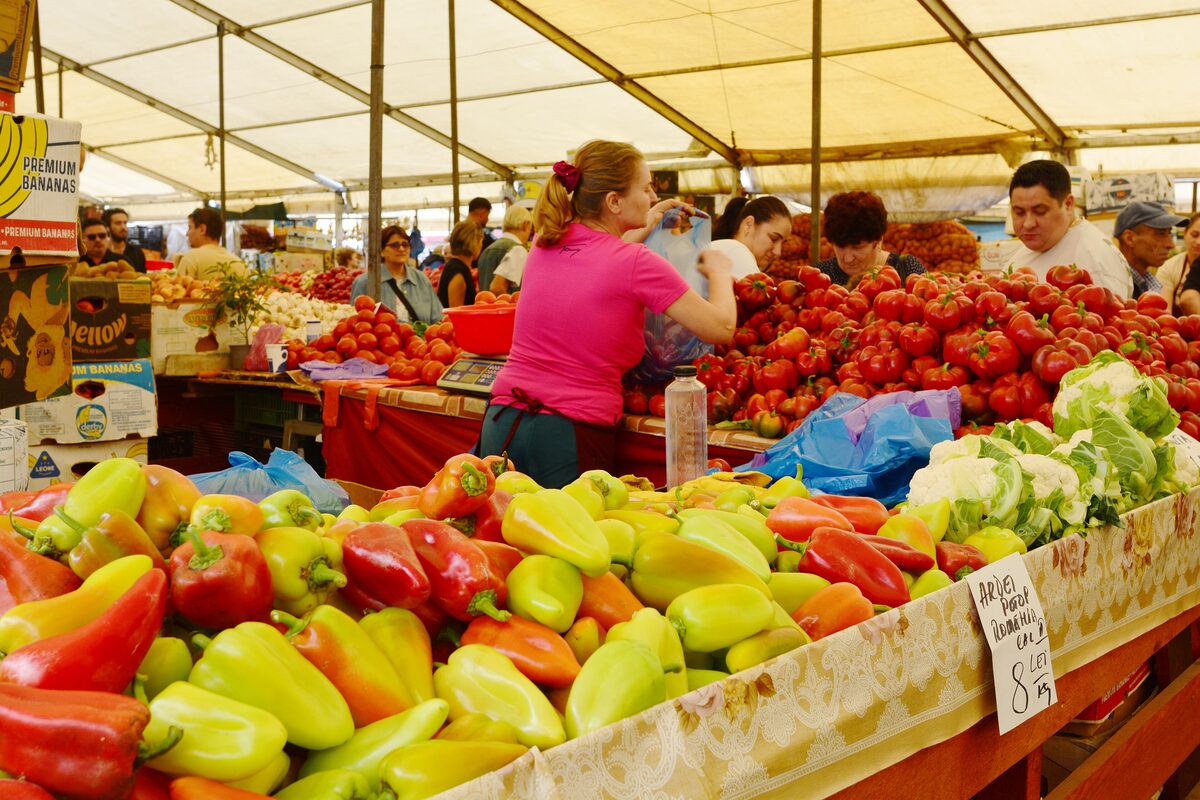 A stall at Obor market piled with red and yellow peppers, tomatoes and cauliflower under a canvas roof, with a handwritten price sign