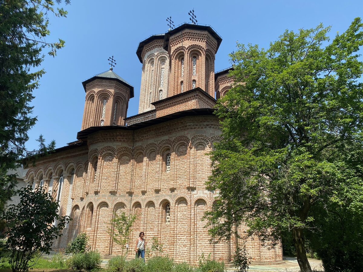 The brick-and-stone church of Snagov Monastery, with its three tall towers and rows of blind arches, among green trees