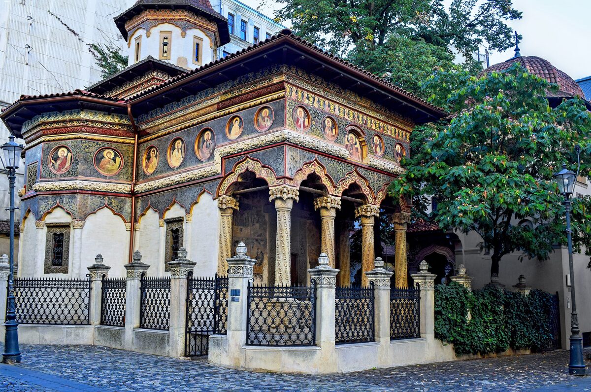 Stavropoleos Church with its painted saints in round medallions and the arched porch on carved stone columns, behind a wrought-iron fence on a cobbled lane