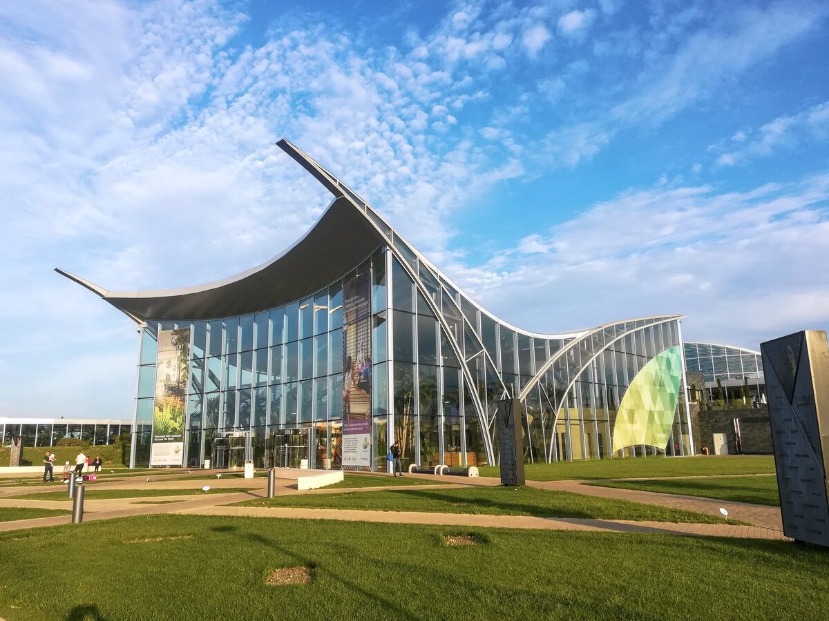 The glass entrance building of Therme București, with its sweeping wave-shaped roof, behind lawns under a blue sky