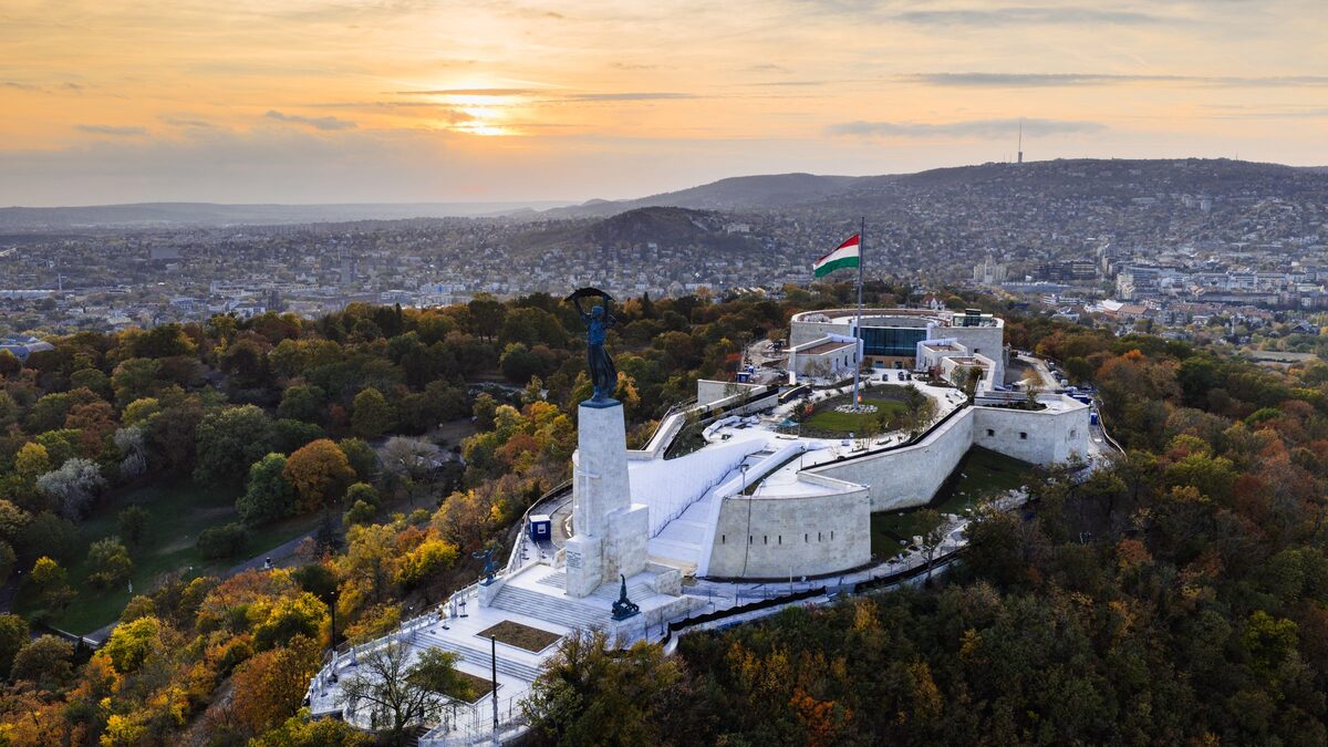 Aerial view of the Liberty Statue and the white walls of the Citadella on Gellért Hill among autumn trees, with Buda behind at sunset