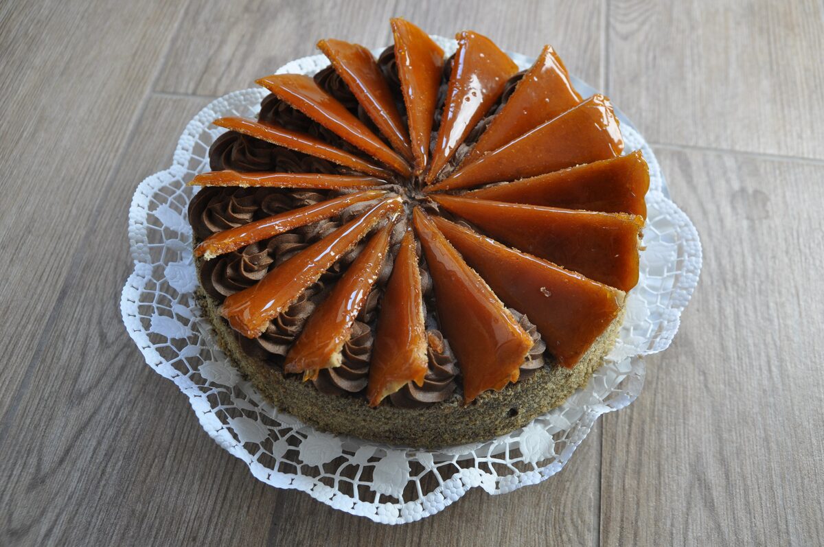 A whole Dobos torte with a ring of shiny caramel-glazed wedges on top, on a lace doily