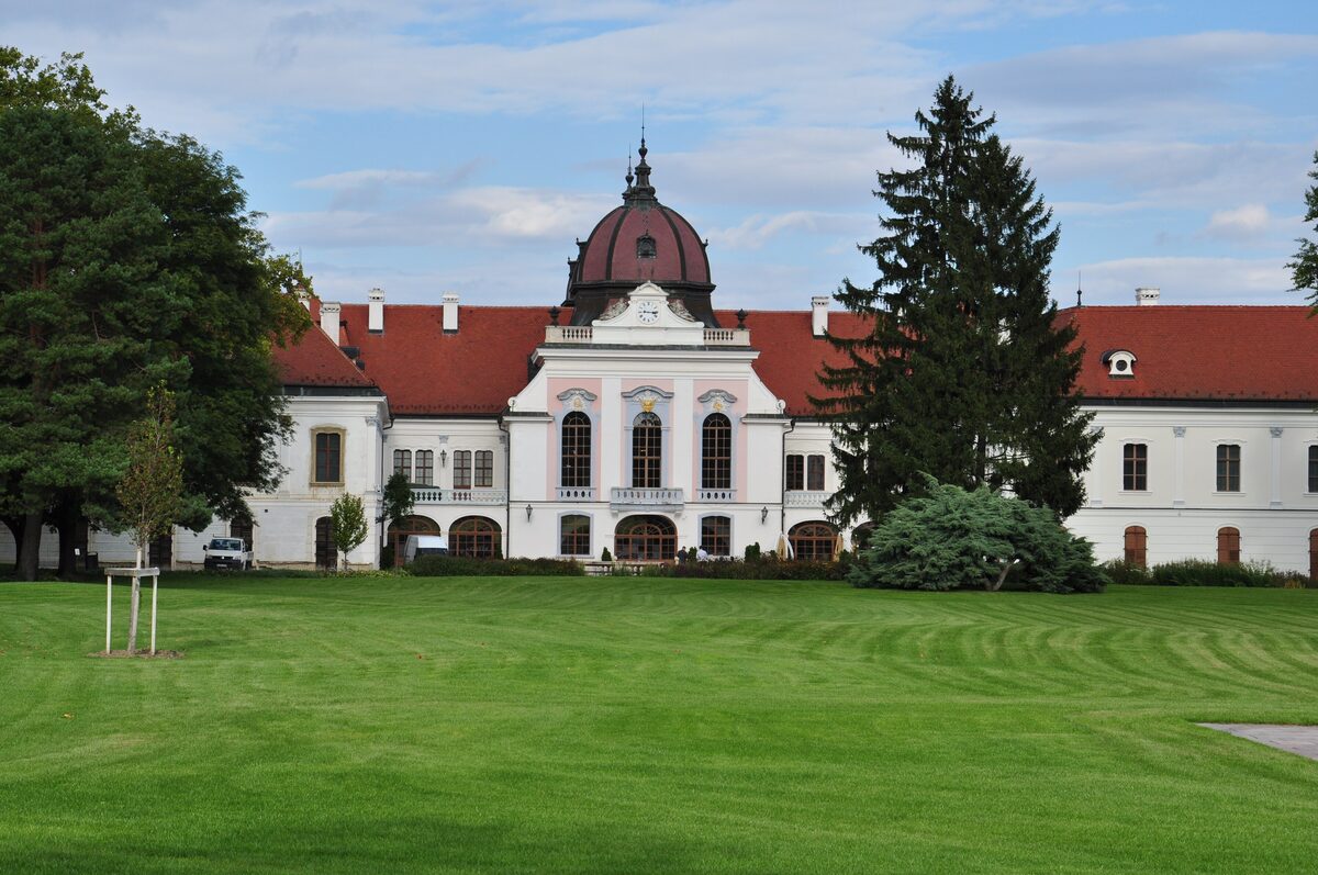 The white Gödöllő Royal Palace with red roofs and a domed center, seen across a wide striped lawn