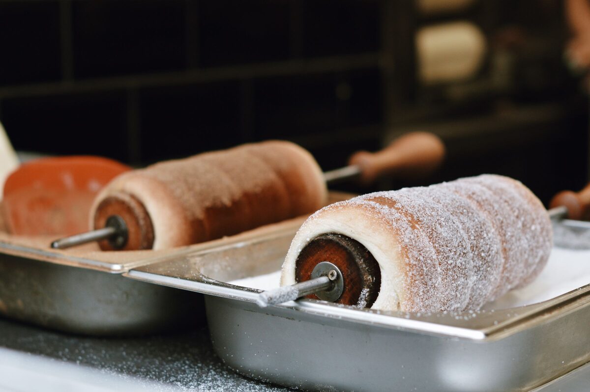 Two chimney cakes coated in sugar on their wooden rollers over a grill