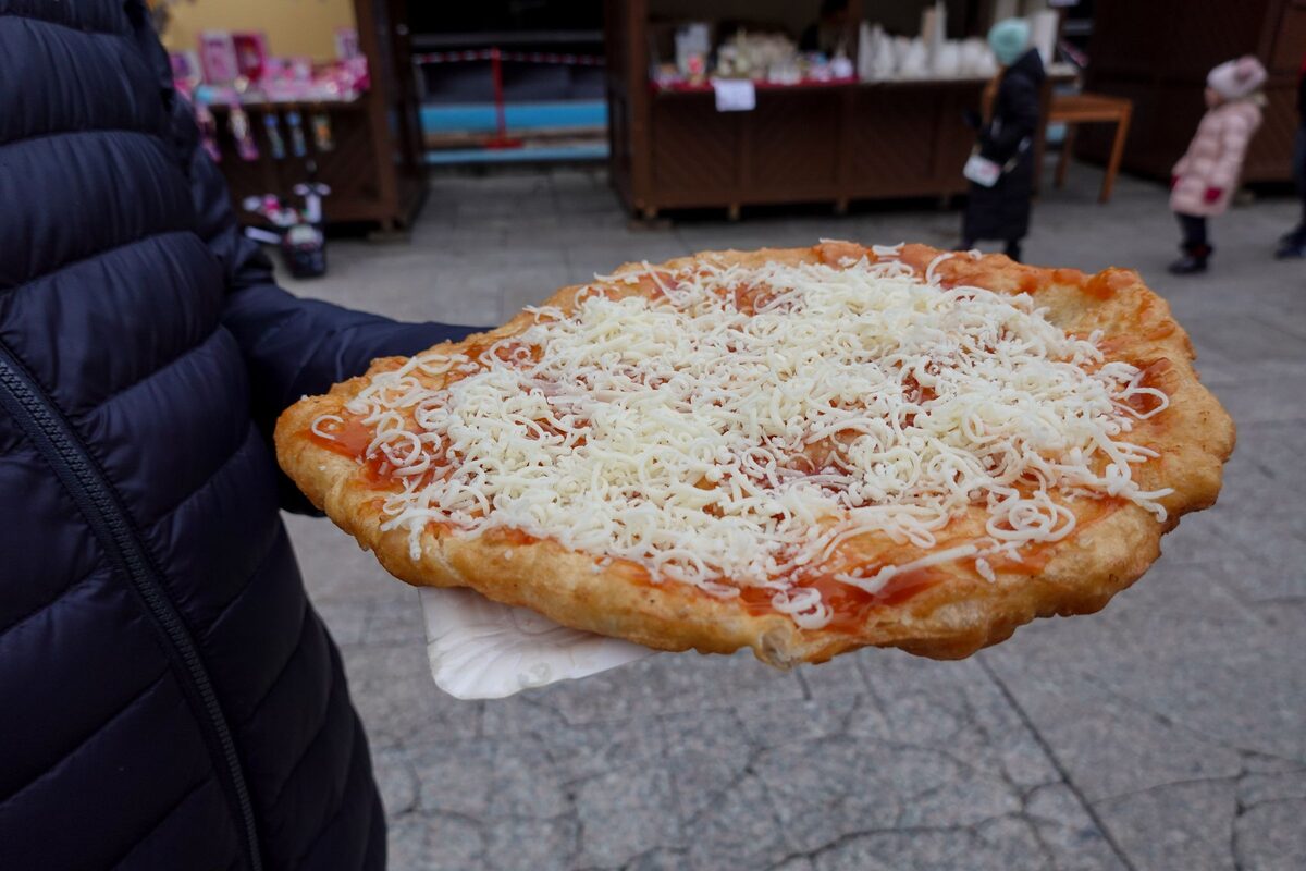 A hand holding a large lángos topped with grated cheese and tomato sauce at an outdoor market