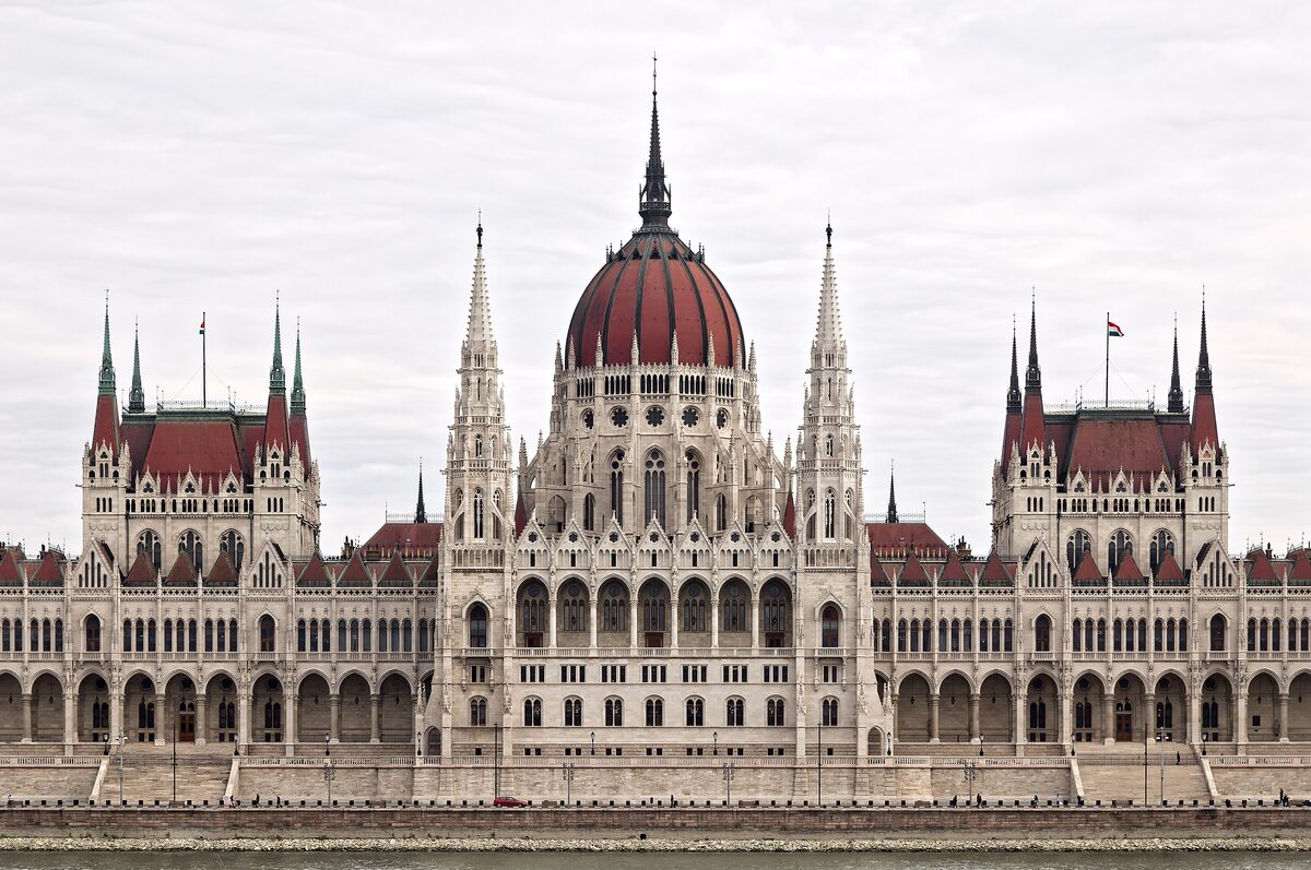 The neo-Gothic Hungarian Parliament with its red dome and spires, seen head-on across the Danube under a pale sky