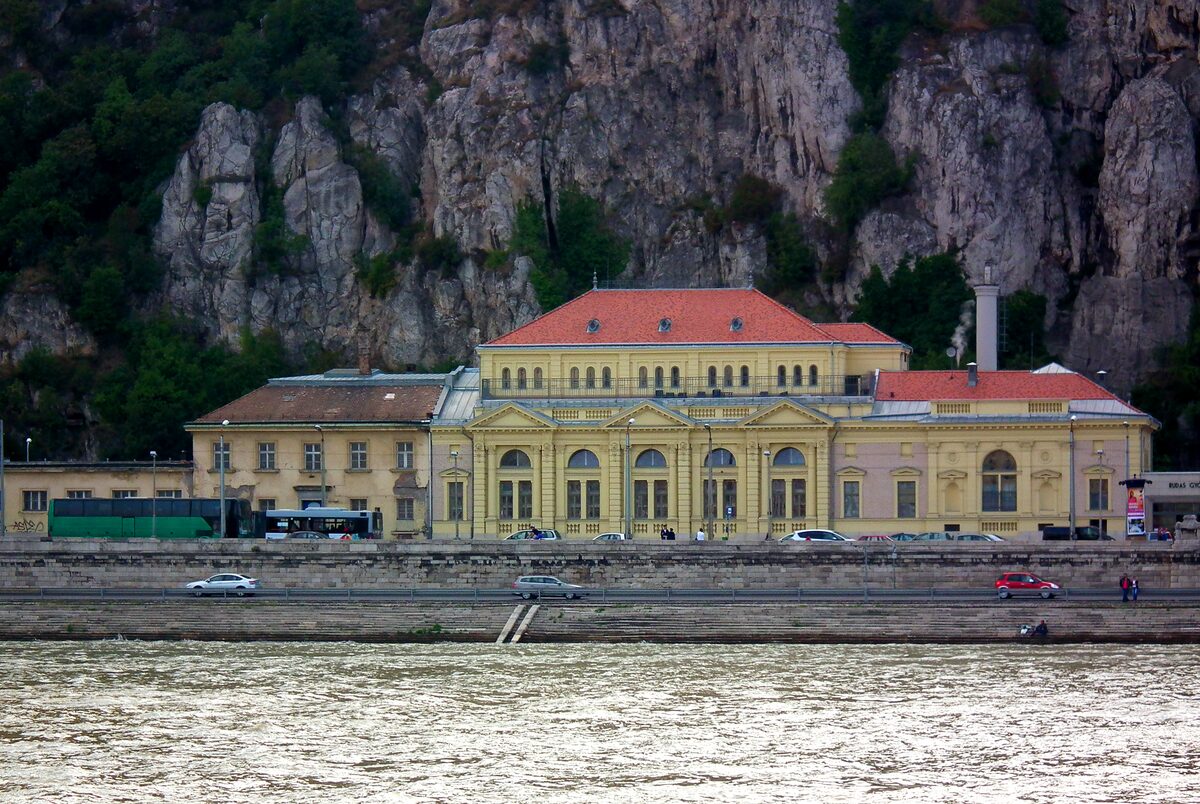 The yellow facade and red roofs of Rudas Bath on the Danube embankment, below the limestone cliffs of Gellért Hill