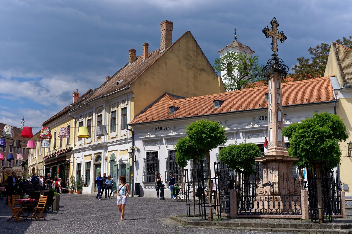 Fő tér in Szentendre, with baroque houses, colorful lampshades strung over a lane and the iron-railed Serbian merchants' cross under a cloudy sky