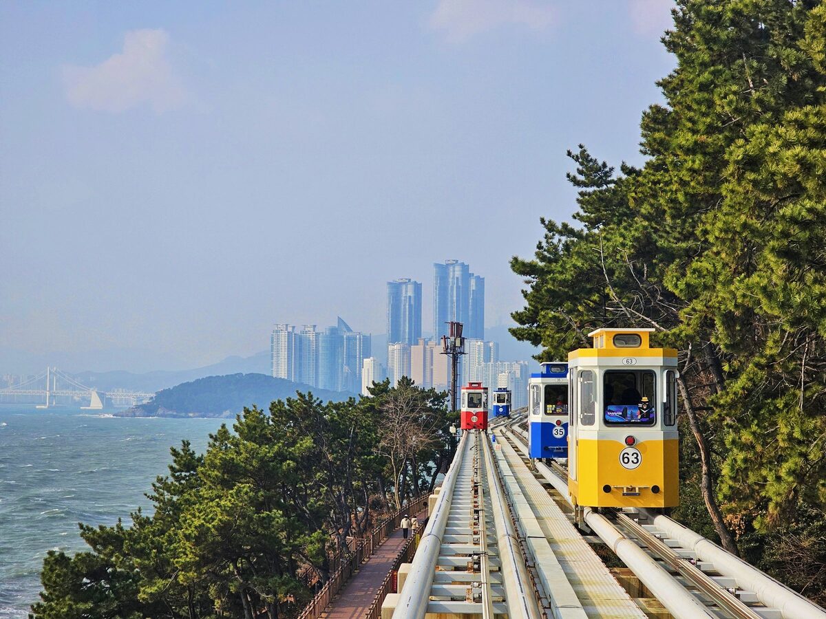 Yellow, blue and red Beach Train cars on the Blueline Park track above the sea, with the Haeundae towers in the haze behind