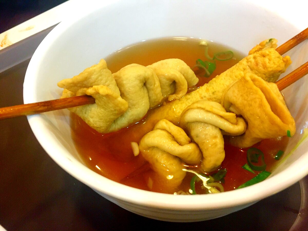 Fish-cake skewers folded onto wooden sticks and served in a white bowl of hot broth with green onion