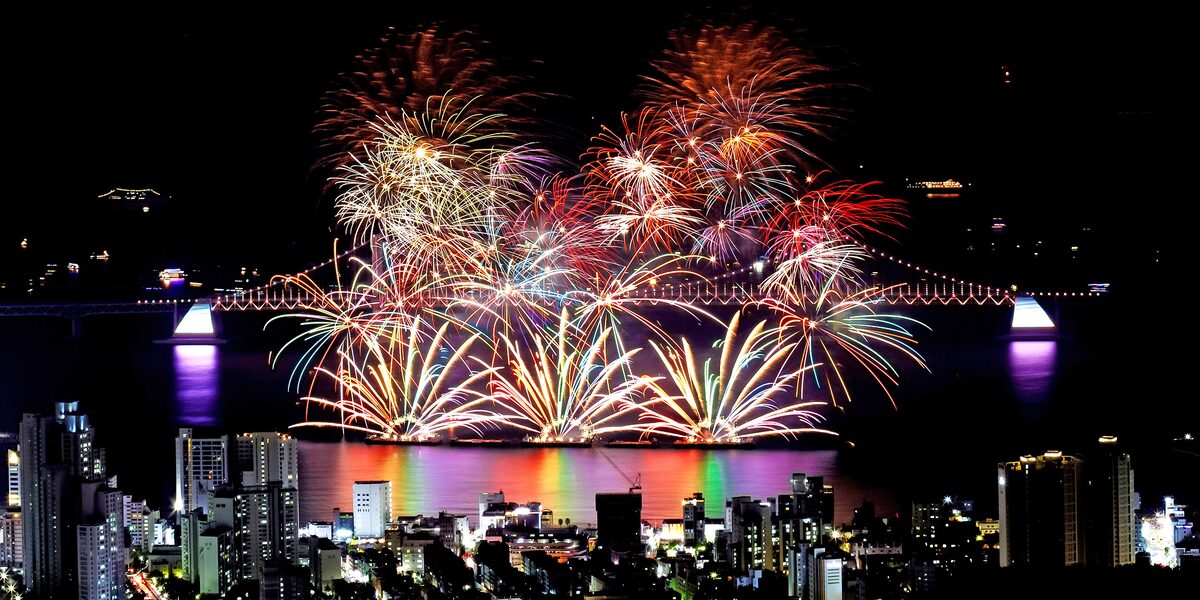 Fireworks bursting in red and white over the water beside a lit bridge, with the city at night