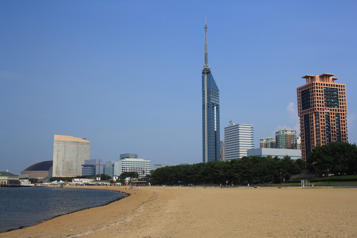 The mirror-glass Fukuoka Tower rising over the sand and pines of Momochi beach under a blue sky