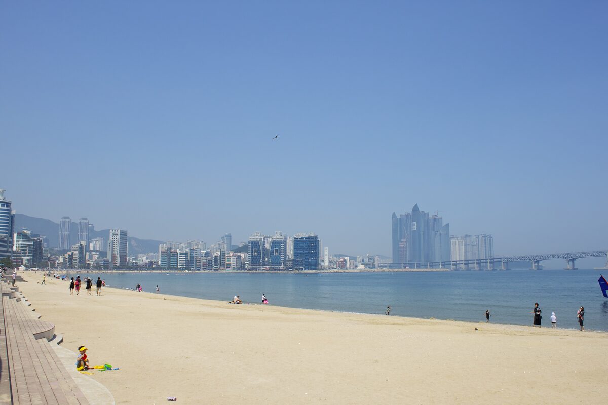 Gwangalli Beach in daylight, with a sandy shore, the beach road and high-rise towers across the bay