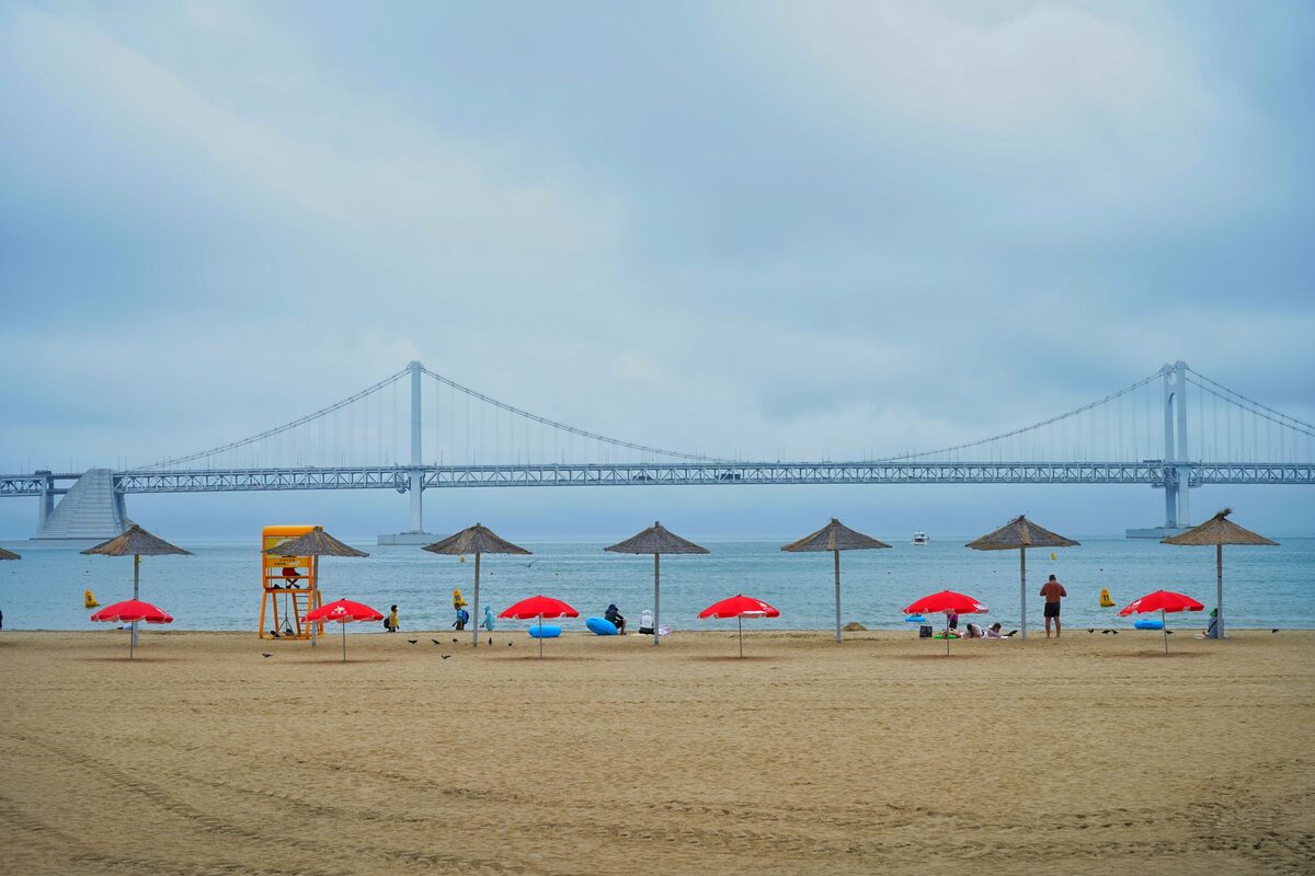 Gwangalli Beach by day, with straw and red beach umbrellas along the sand and the long Gwangan Bridge across the bay