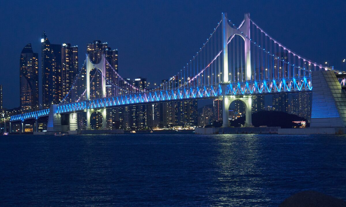 The Gwangan Bridge lit up at night in blue, with high-rise towers glowing on the far side of the bay