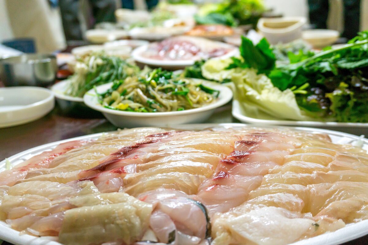 Thin slices of raw fish arranged on a plate, beside a spread of lettuce and leafy greens