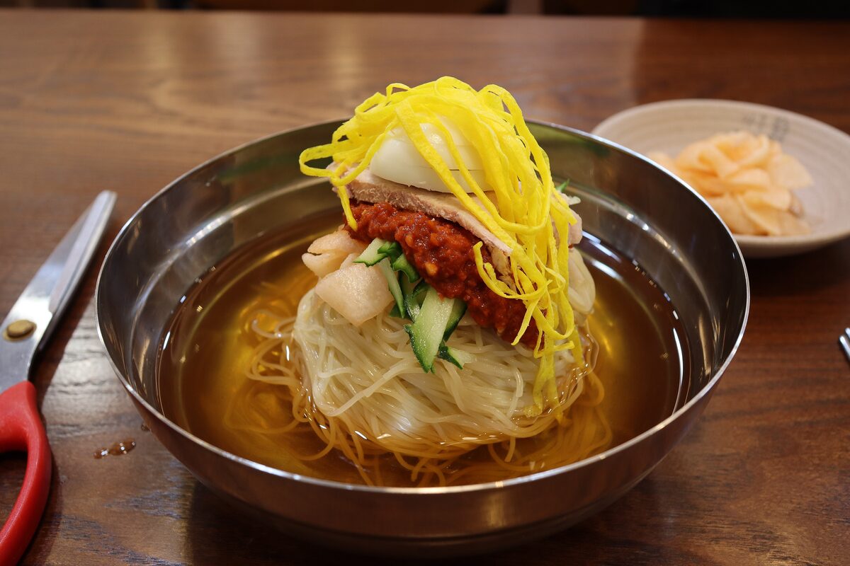 A steel bowl of cold milmyeon noodles with a mound of red chili sauce and yellow strips on top