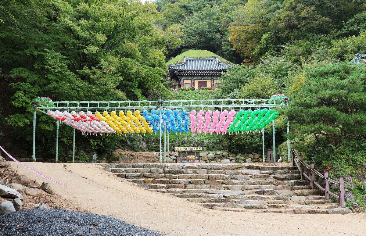 Stone steps climbing a wooded hill under a row of colored lanterns, toward the Seokguram Grotto