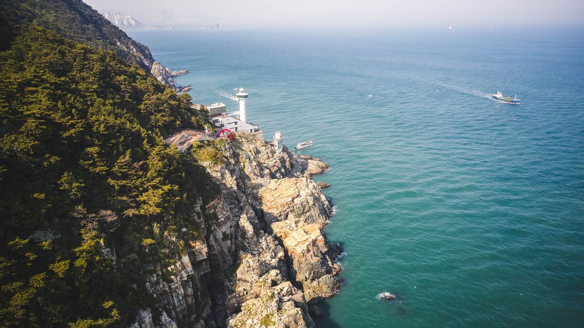 Pine-covered rock cliffs of Taejongdae dropping into turquoise water, with a white lighthouse on the point