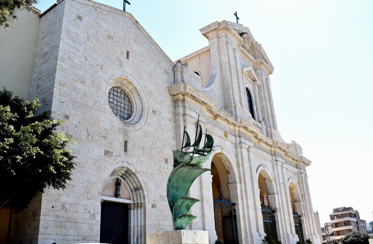 The white limestone facades of the old Bonaria sanctuary and the newer basilica beside it, with a bronze ship sculpture in front