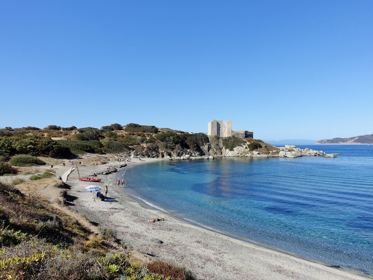 A small grey-sand beach on a bay of clear blue water, with the Fortezza Vecchia tower on the rocky point beyond