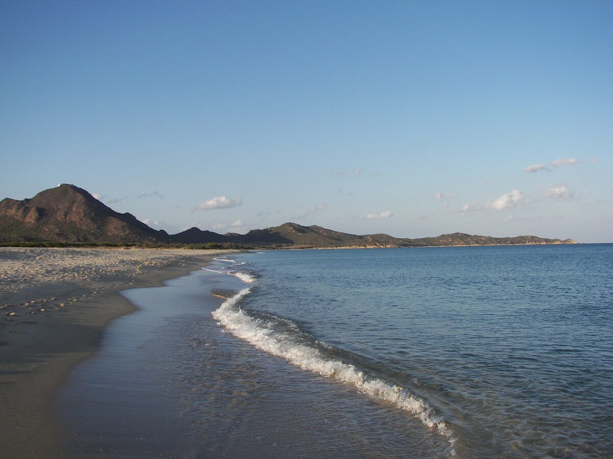 A wave breaking on the long sand beach of Costa Rei, with mountains at the end of the bay