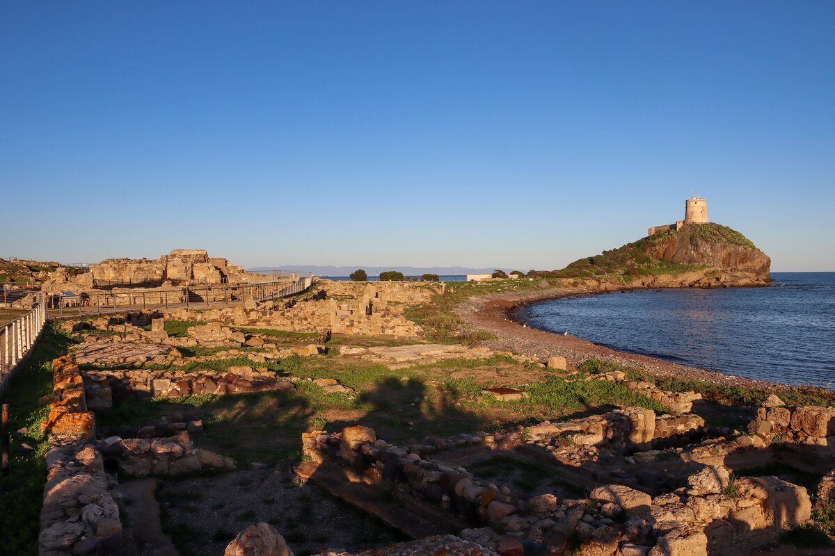 The excavated Roman streets and walls of Nora at golden hour, with the Torre del Coltellazzo on the headland beside the sea