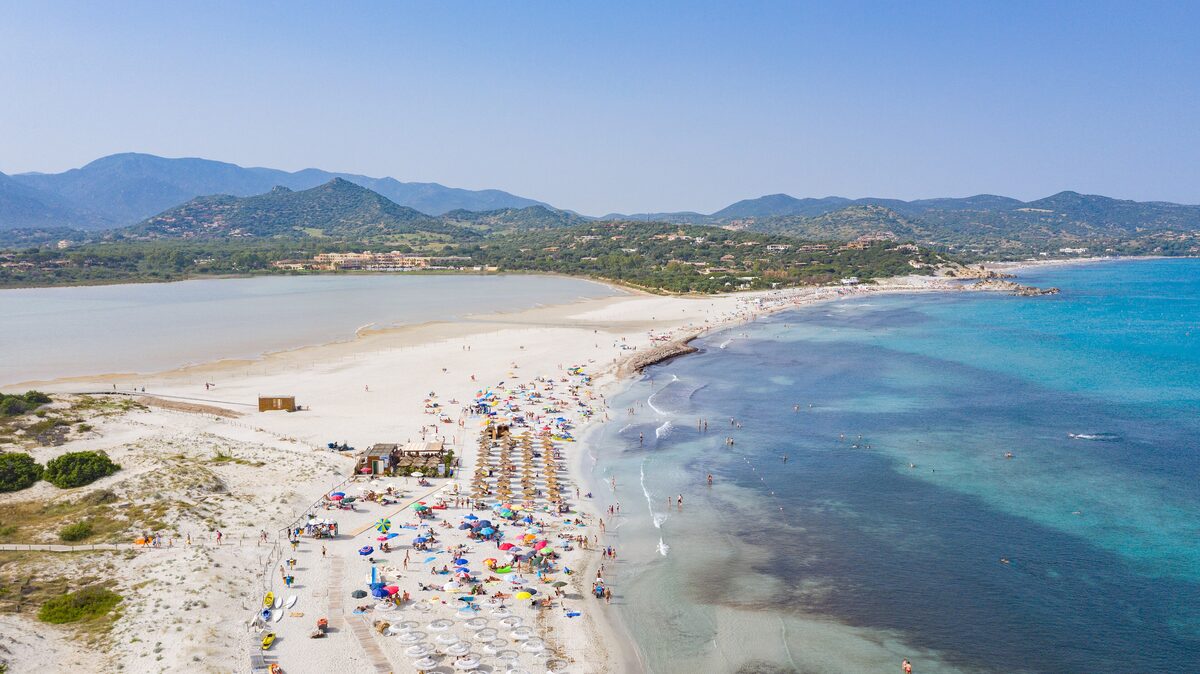 Aerial view of the white sand of Porto Giunco beach with umbrellas, the turquoise sea on one side and the Notteri pond on the other