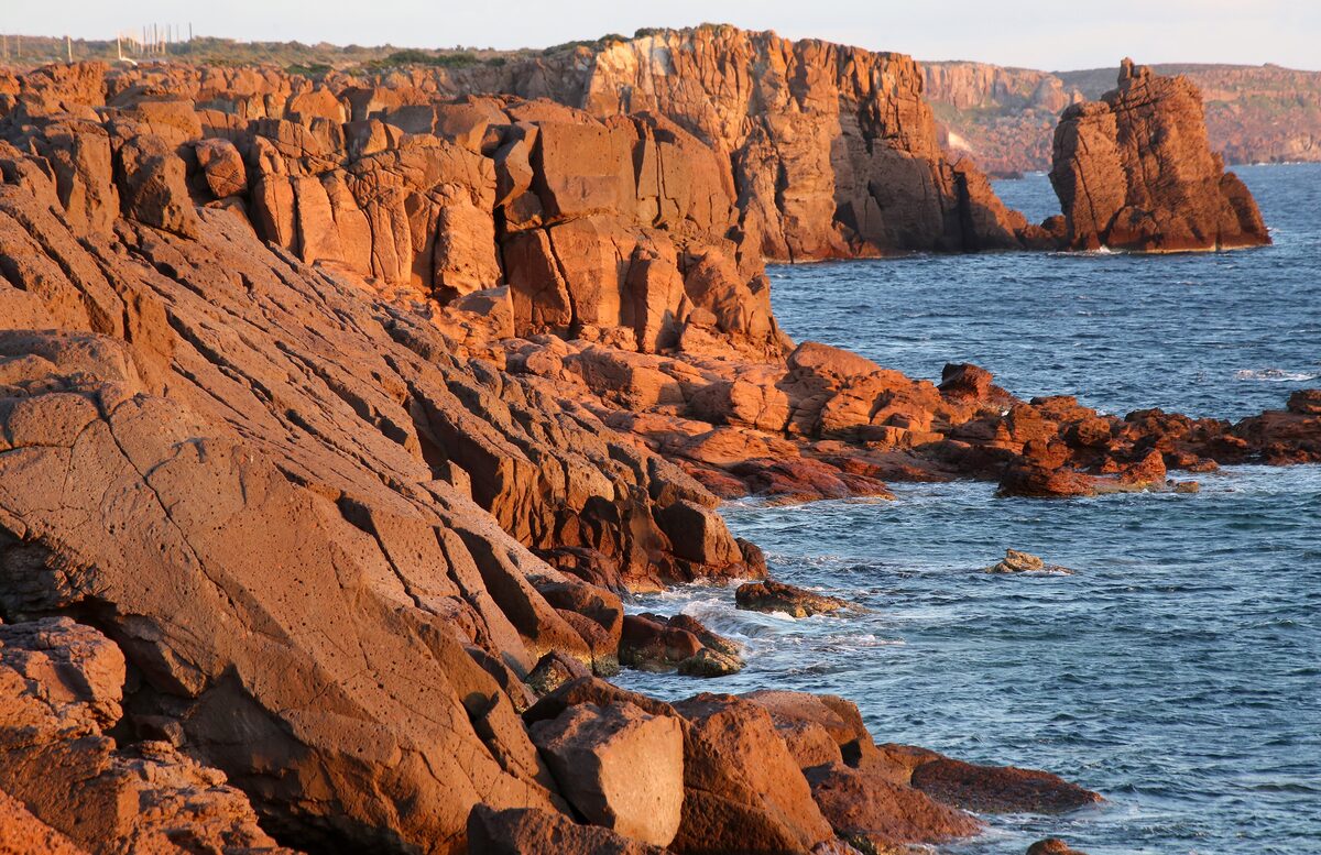 Red volcanic cliffs and sea stacks on the coast of Sant'Antioco island, lit by low sun