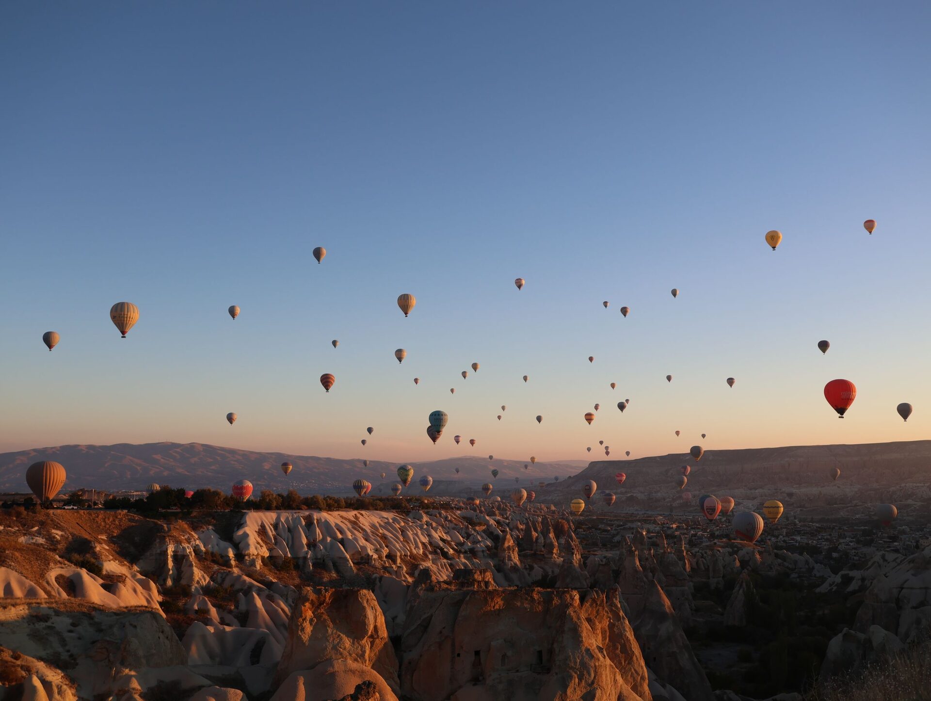 Dozens of hot-air balloons in a clear dawn sky over the fairy chimneys of Göreme, lit orange by the rising sun