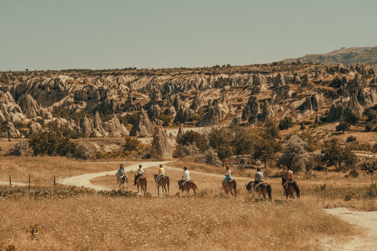 A line of riders on horseback crossing dry golden grassland towards Cappadocia's rock formations