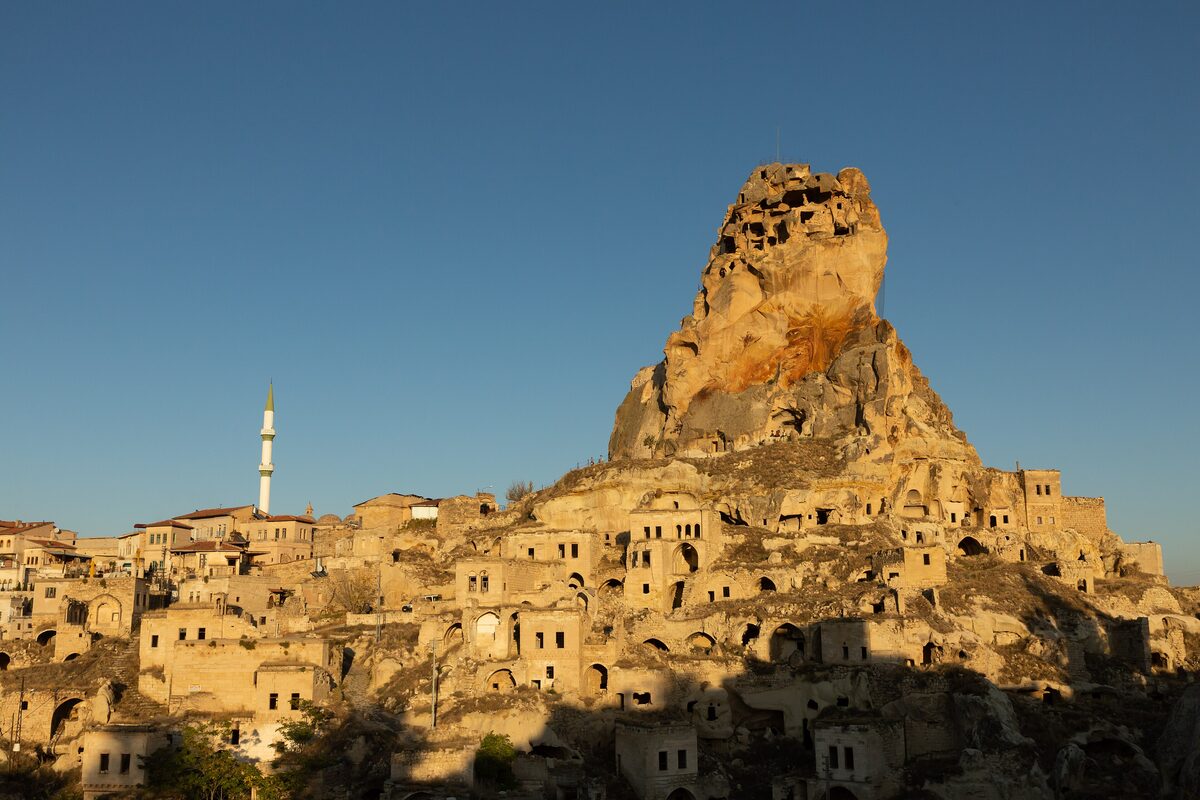 The castle rock and cave houses of Ortahisar in low golden sun, with a minaret on the left