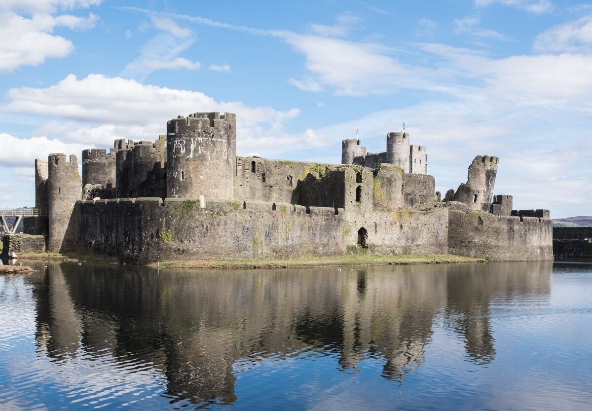 Caerphilly Castle, gray stone towers and walls rising from its lake, reflected in still water under a blue sky