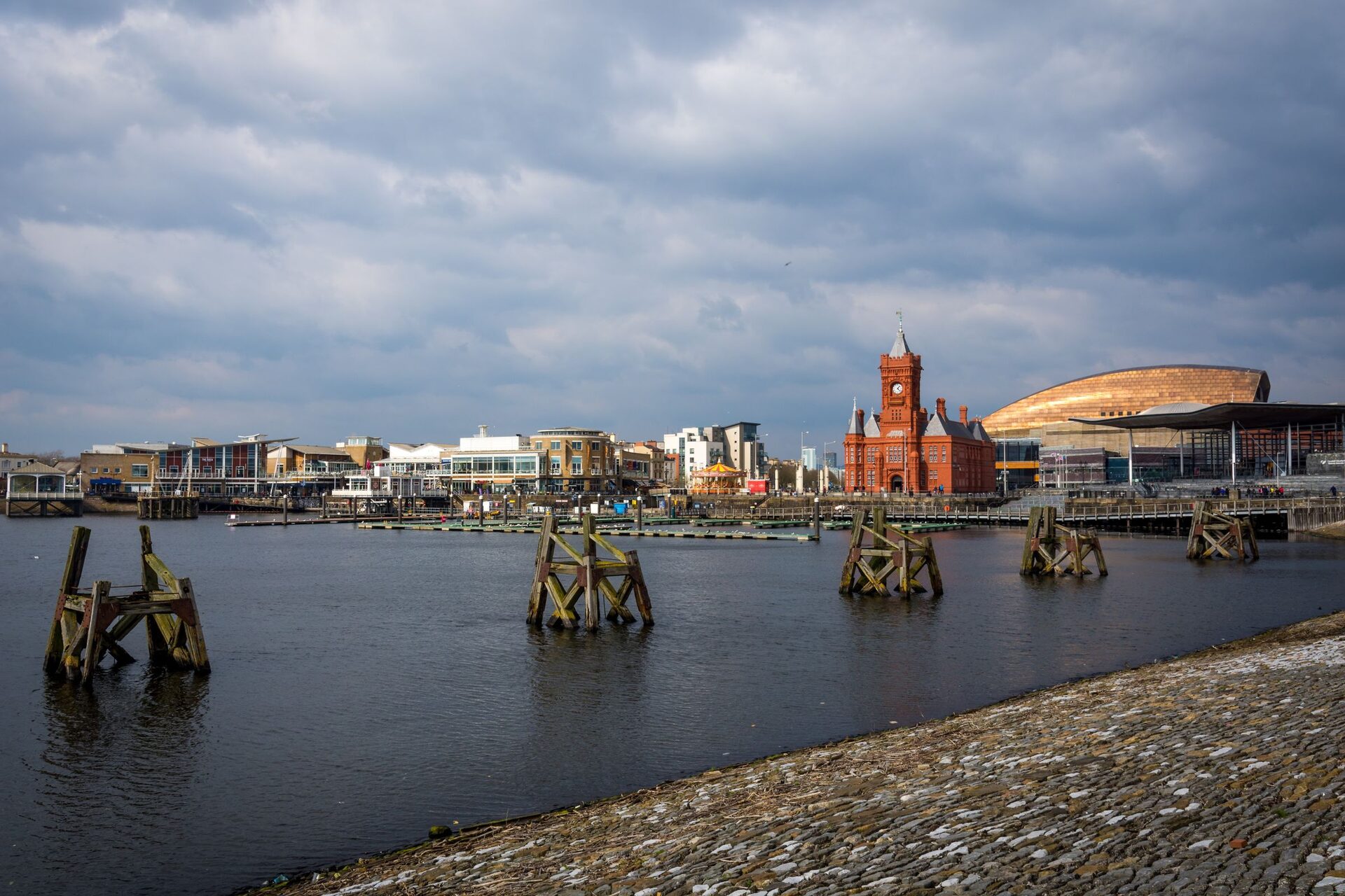 Cardiff Bay under a cloudy sky, with old wooden posts in the water, the red brick Pierhead clock tower and the bronze roof of the Wales Millennium Centre