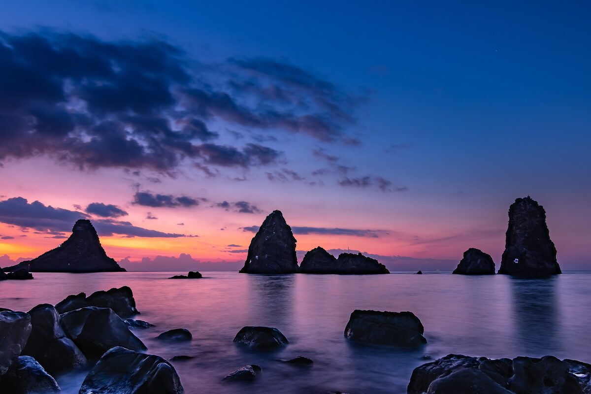 Dark rocks and sea stacks off Aci Trezza at sunrise, with a purple and pink sky over the water