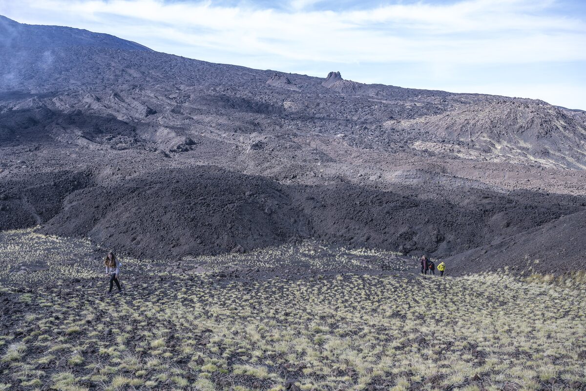 Dark lava slopes and cinder fields high on Mount Etna, with a few hikers and a hazy sky over the plain