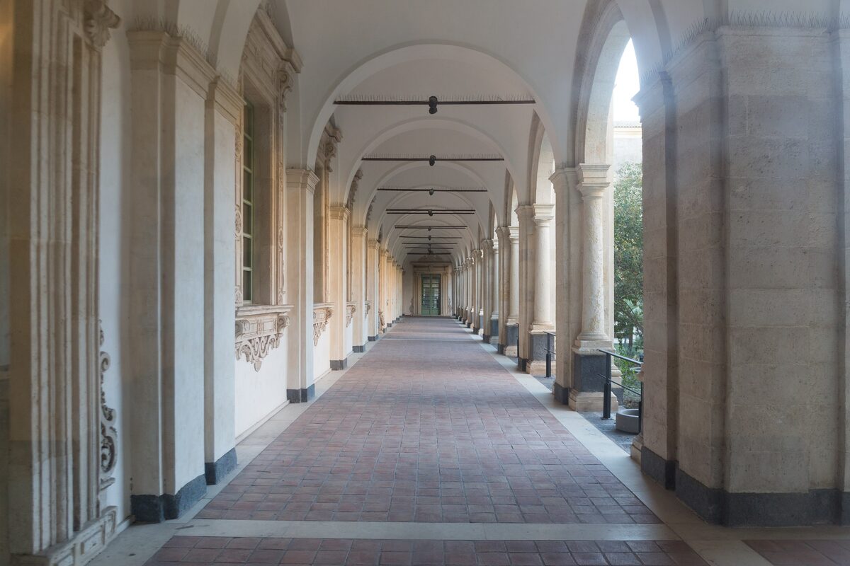 Long arcaded corridor with white columns and a tiled floor inside the Benedictine monastery of San Nicolo l'Arena in Catania